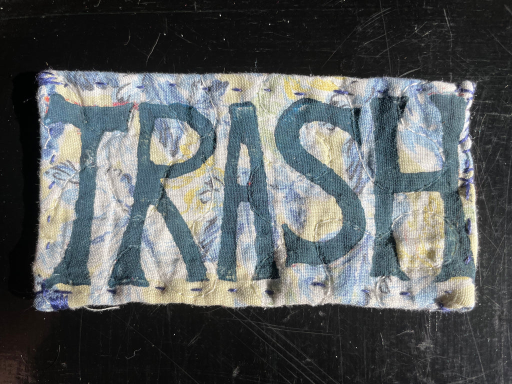 Patch with 'TRASH' printed on a quilted  green and blue fabric, placed on a dark surface.