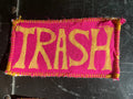 Fabric patch with 'TRASH' text on a pink background, attached to a black surface.
