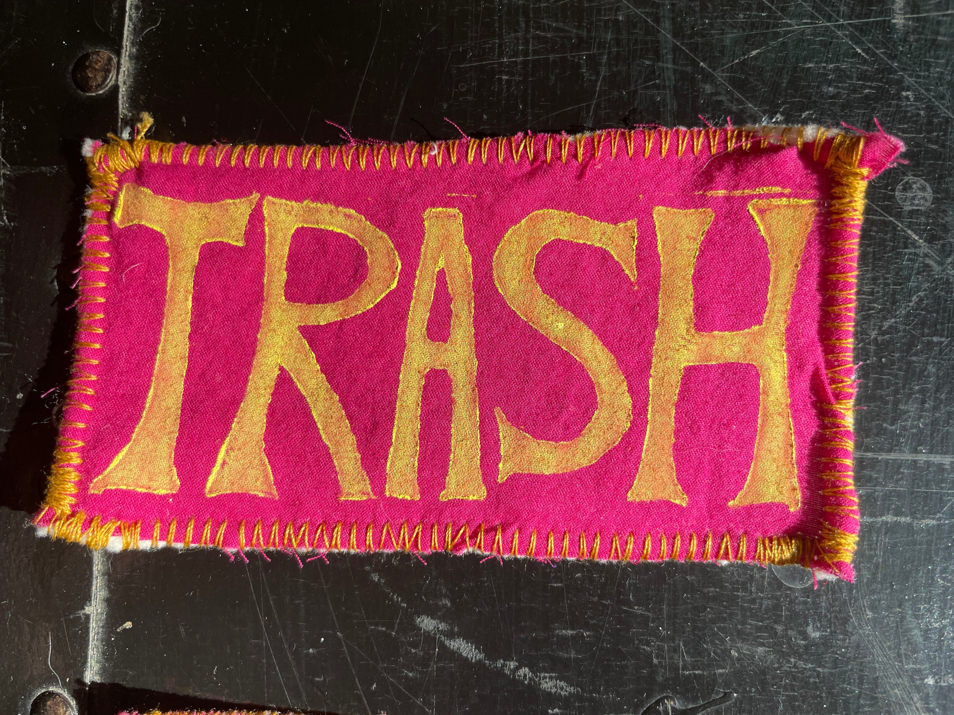 Fabric patch with 'TRASH' text on a pink background, attached to a black surface.