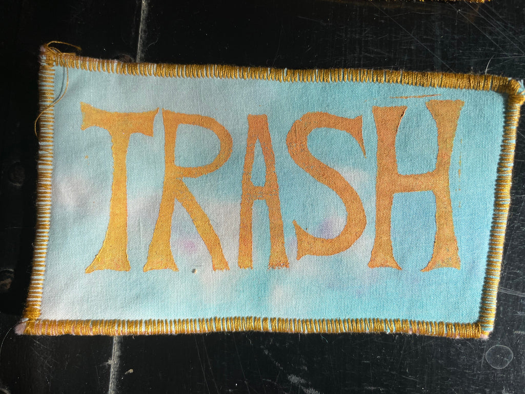 Fabric patch with 'TRASH' printed in gold on a light blue tie dye background