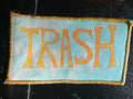 Fabric patch with 'TRASH' printed in gold on a light blue tie dye background